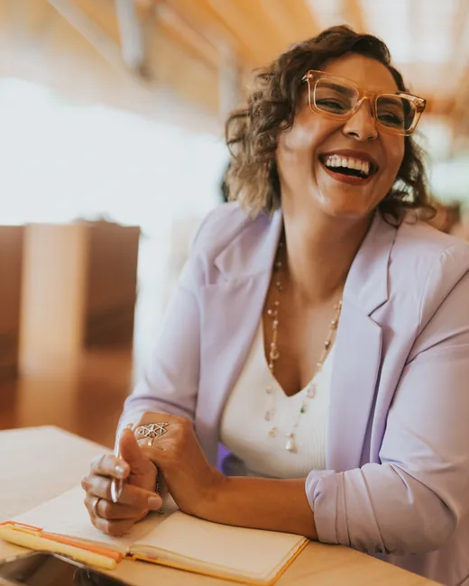 Ju Bella rindo, sentada à mesa com um caderno aberto